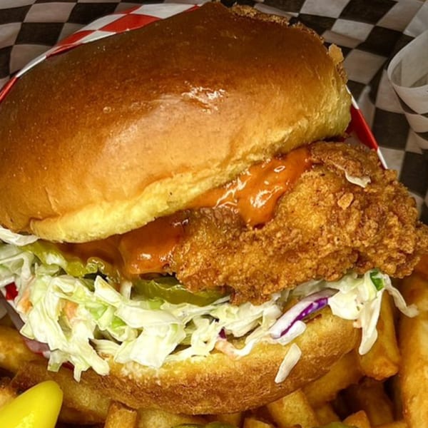 Krispi Hot Chicken sandwich with sauce, coleslaw, and pickles, served with fries in a basket.