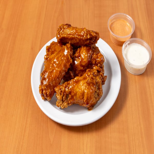 A white plate holds several glossy, brown chicken wings with two dipping sauces on a wooden table.