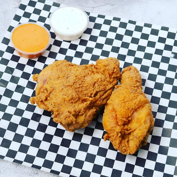 Two pieces of Krispi Hot Chicken fried chicken with two dipping sauces on checkered paper.